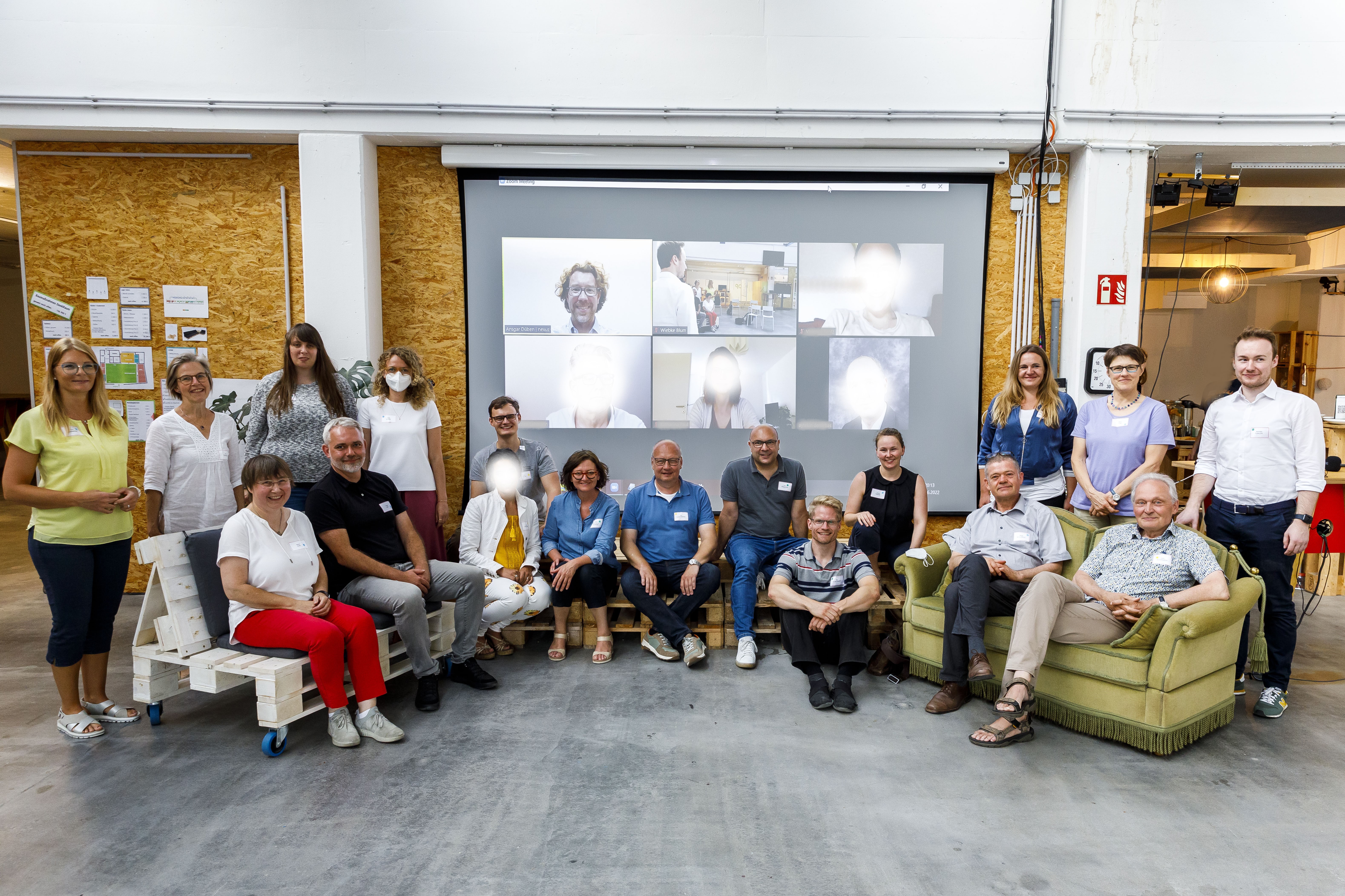 Mitglieder sitzen und stehen im Halbkreis, im Hintergrund die Leinwand mit digital Teilnehmenden