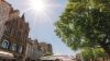 Das Bild zeigt eine Szene auf dem Braunschweiger Kohlmarkt. Die Sonne scheint gleißend vom Himmel, im rechten Bildbereich steht ein großer Baum mit grünem Laub. darunter befinden sich Tische, an denen Menschen sitzen. Diese sind z.T. durch weiße Sonnenschirme vor der Hitze geschützt. Im linken Bildrand befindet sich eine Häuserfassade.